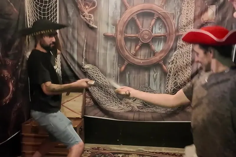 Deux joueurs en chapeau de pirate s'affrontent au sabre devant une barre de navire, dans une salle de Défis Pirate Perpignan