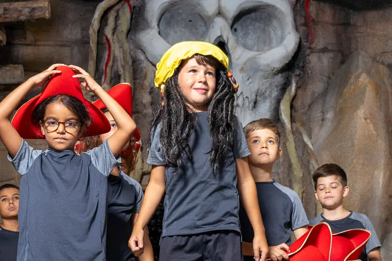Des enfants coiffés de chapeaux de pirate devant la tête de pirate géante de Défis Pirate Perpignan, dont une fillette en perruque et chapeau jaune