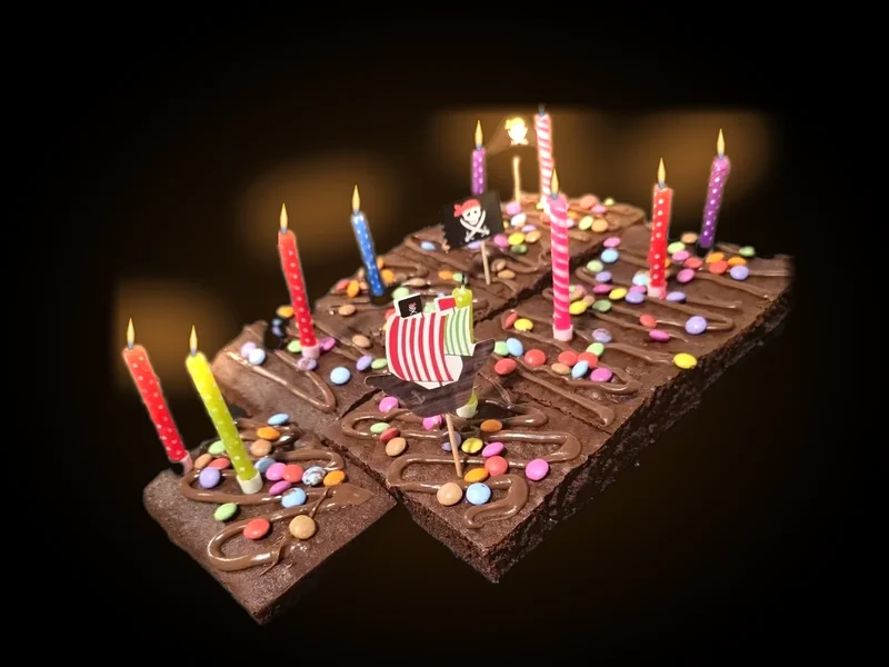 Un gâteau d'anniversaire au chocolat coupé en parts, couvert de bonbons de couleur, avec ses bougies allumées et des décorations pirates, sur fond noir