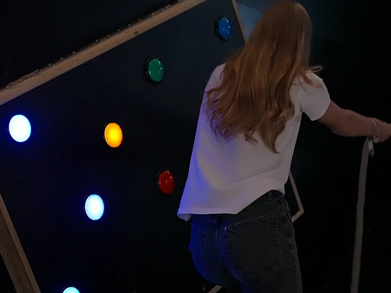 Une joueuse, de dos, face au mur de boutons lumineux de couleur