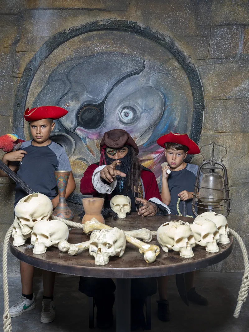 Deux enfants coiffés de chapeaux de pirate entourent un pirate costumé, autour d'une table couverte de crânes