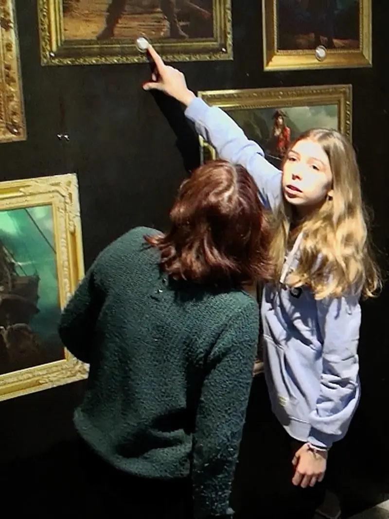 Une jeune joueuse pointe un indice parmi les tableaux de pirates, à côté d'une autre joueuse